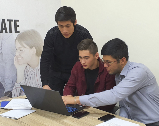 Three male Tashkent students pointing to a laptop screen, while smiling and discussing what they see