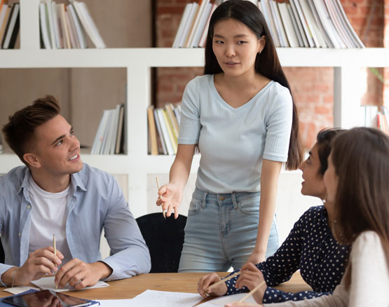Group of students listening as one young woman is speaking in classroom around a table with papers.