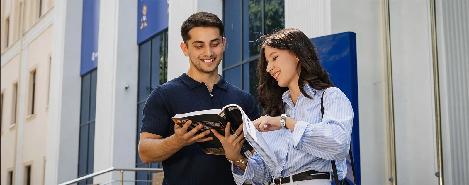 Two students stand in front of Tashkent campus and look at book together.