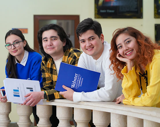 Group of four students holding notebooks lean on balcony railing.